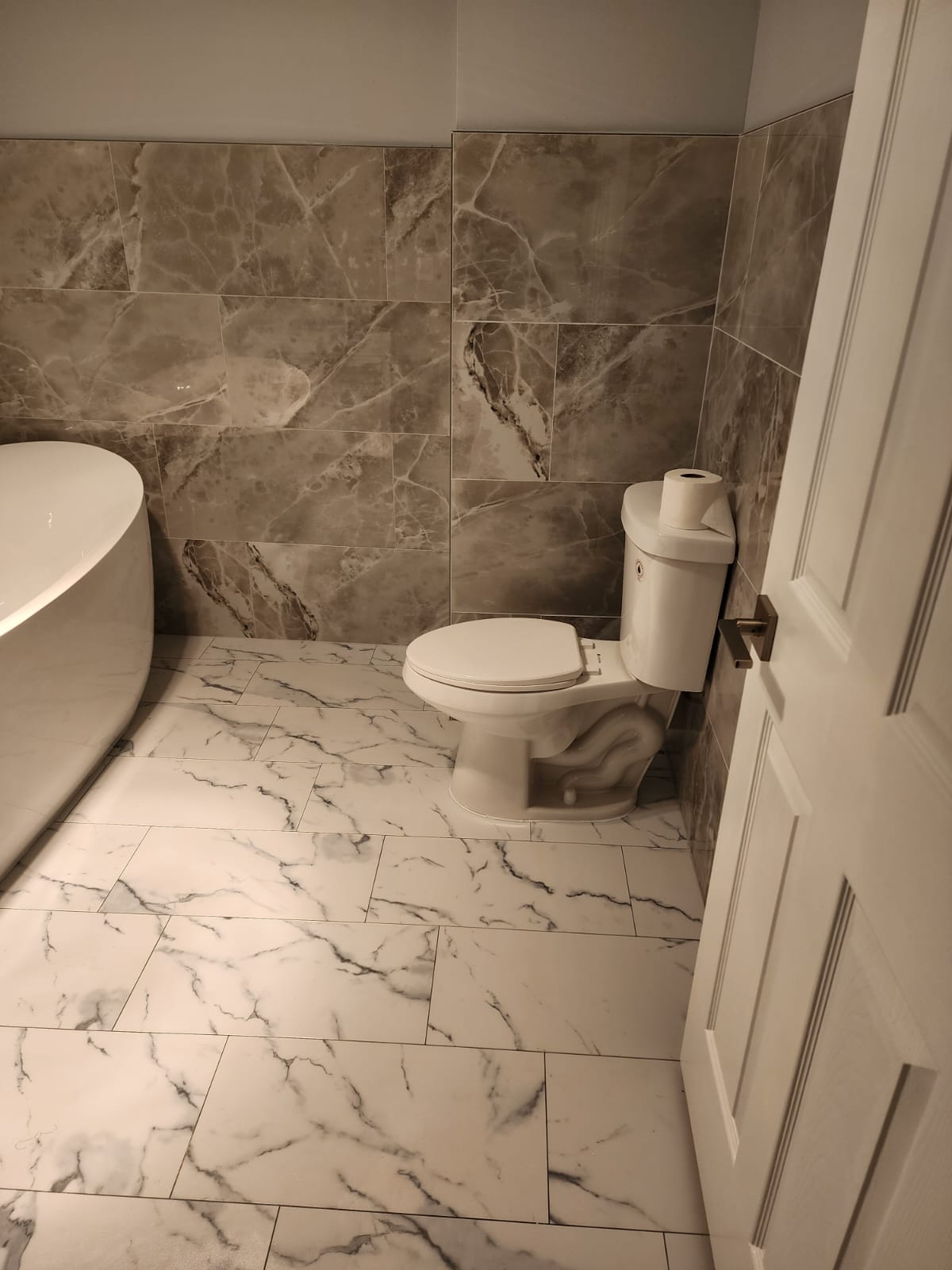 Freestanding soaking tub against brown marble tile with white marble-look floor.
