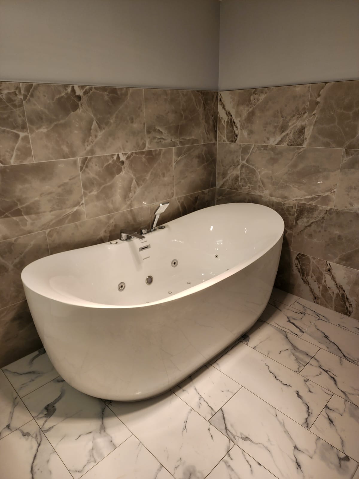 Freestanding jetted tub against brown marble tile with white marble floor.