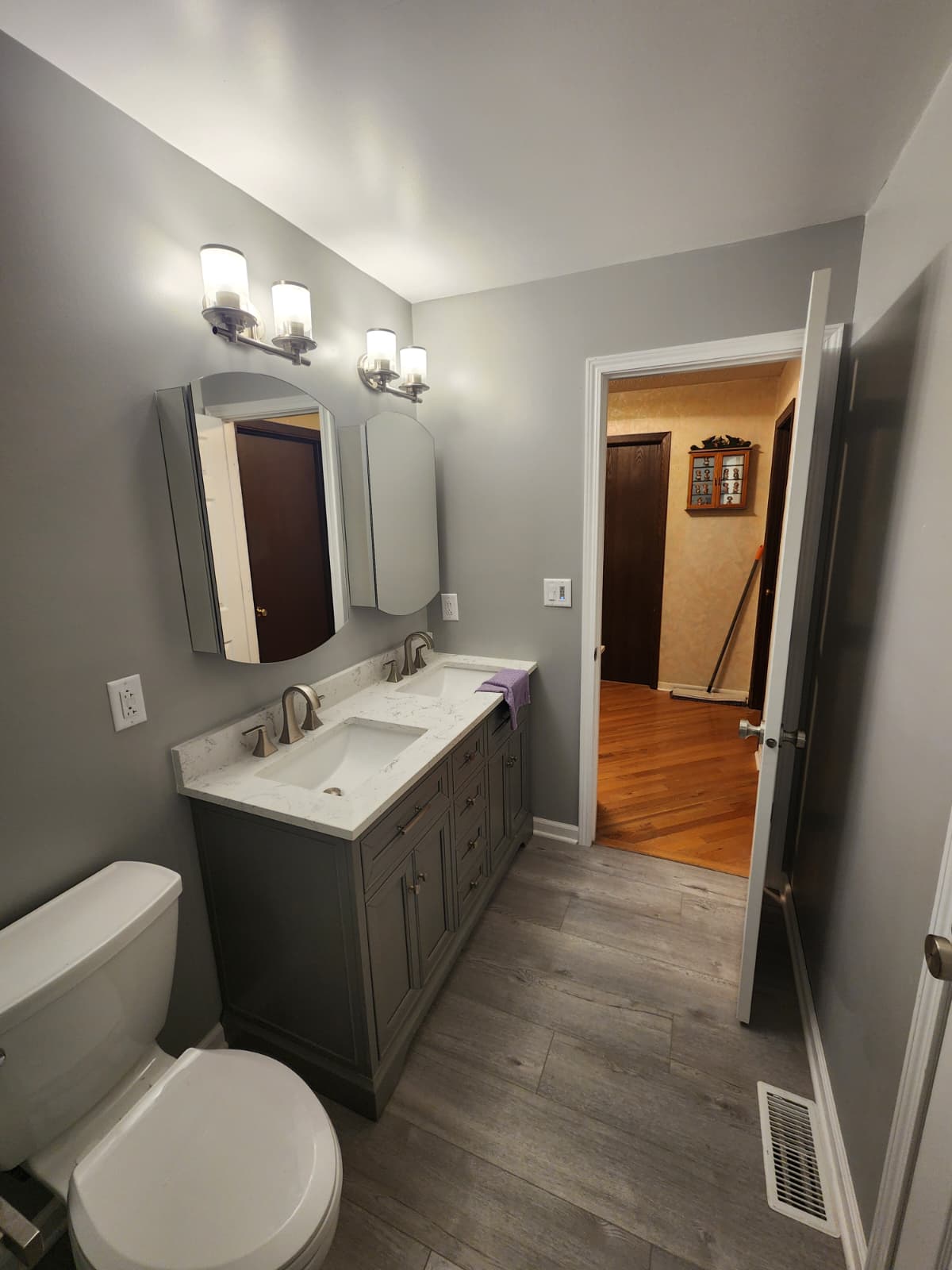 Modern bathroom with gray double vanity, white quartz top, arched mirrors, and wood-look flooring.