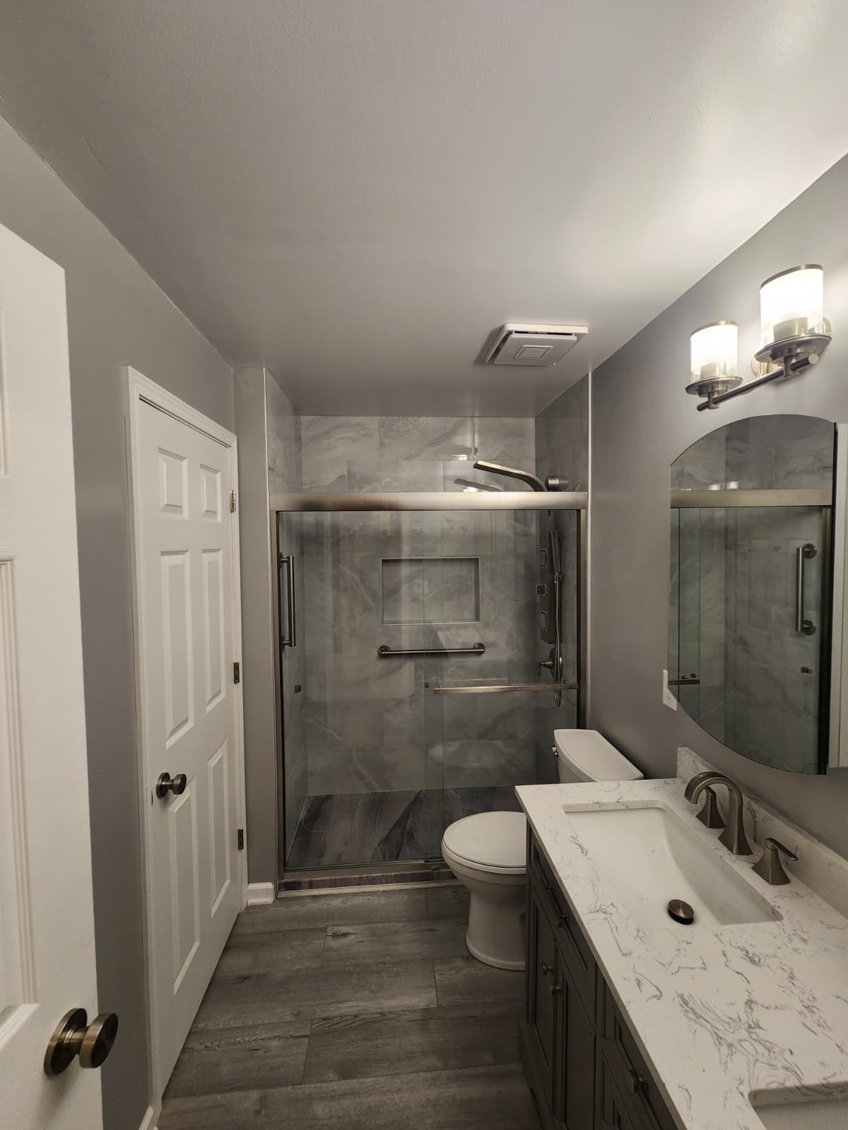 Remodeled bathroom with a gray vanity, marble-look quartz counter, arched mirror, and a sliding-glass marble-tiled shower with grab bar.