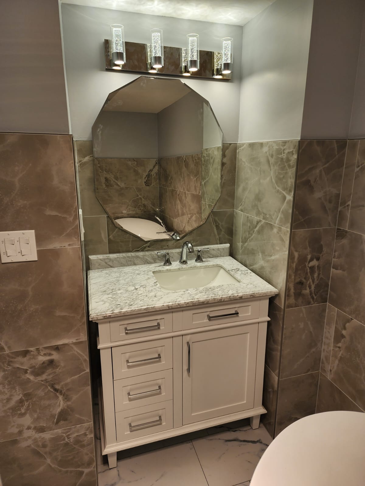 White vanity with octagonal beveled mirror and bubble vanity lights against brown marble tile.