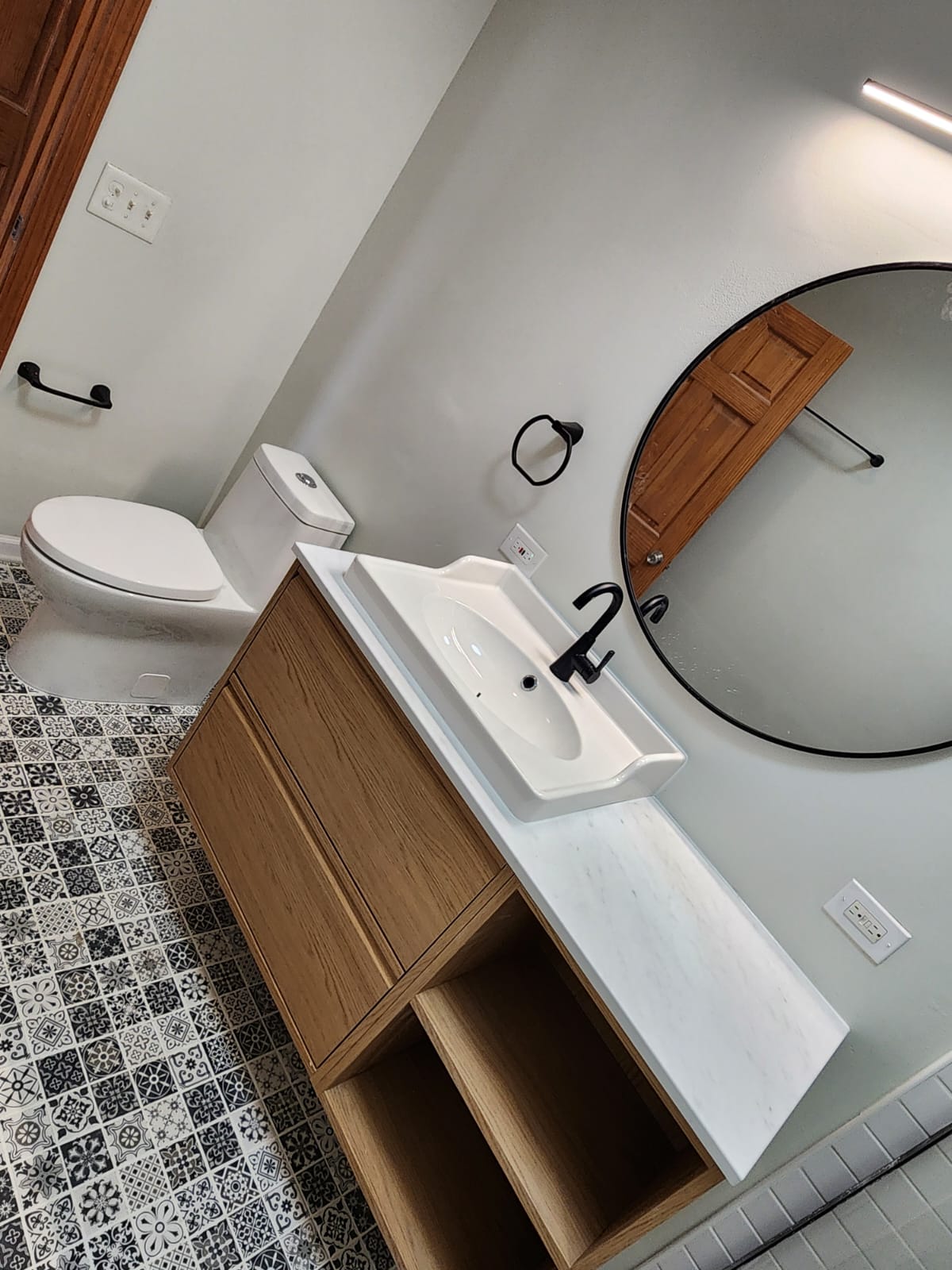 Floating wood vanity with white sink and round mirror above patterned cement floor tile.