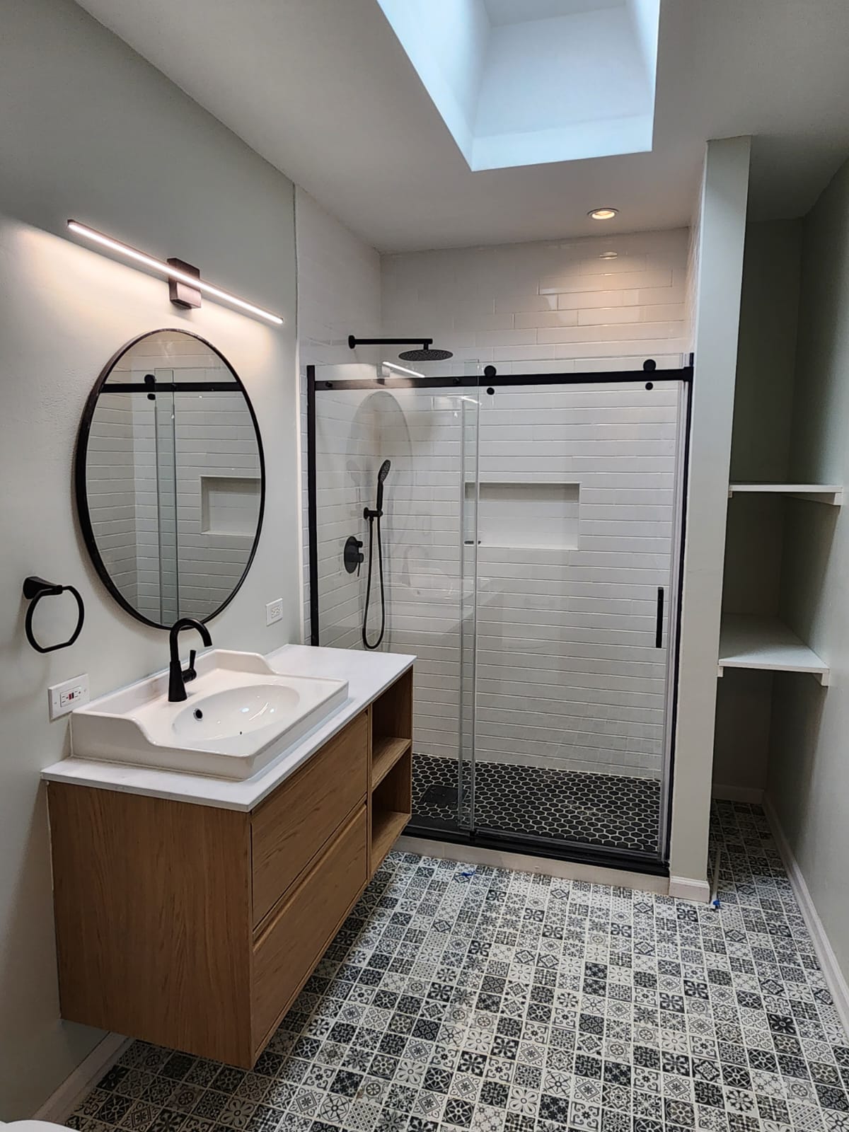 Remodeled bathroom with floating wood vanity, round black-framed mirror, and a glass walk-in shower with subway tile beneath a skylight.