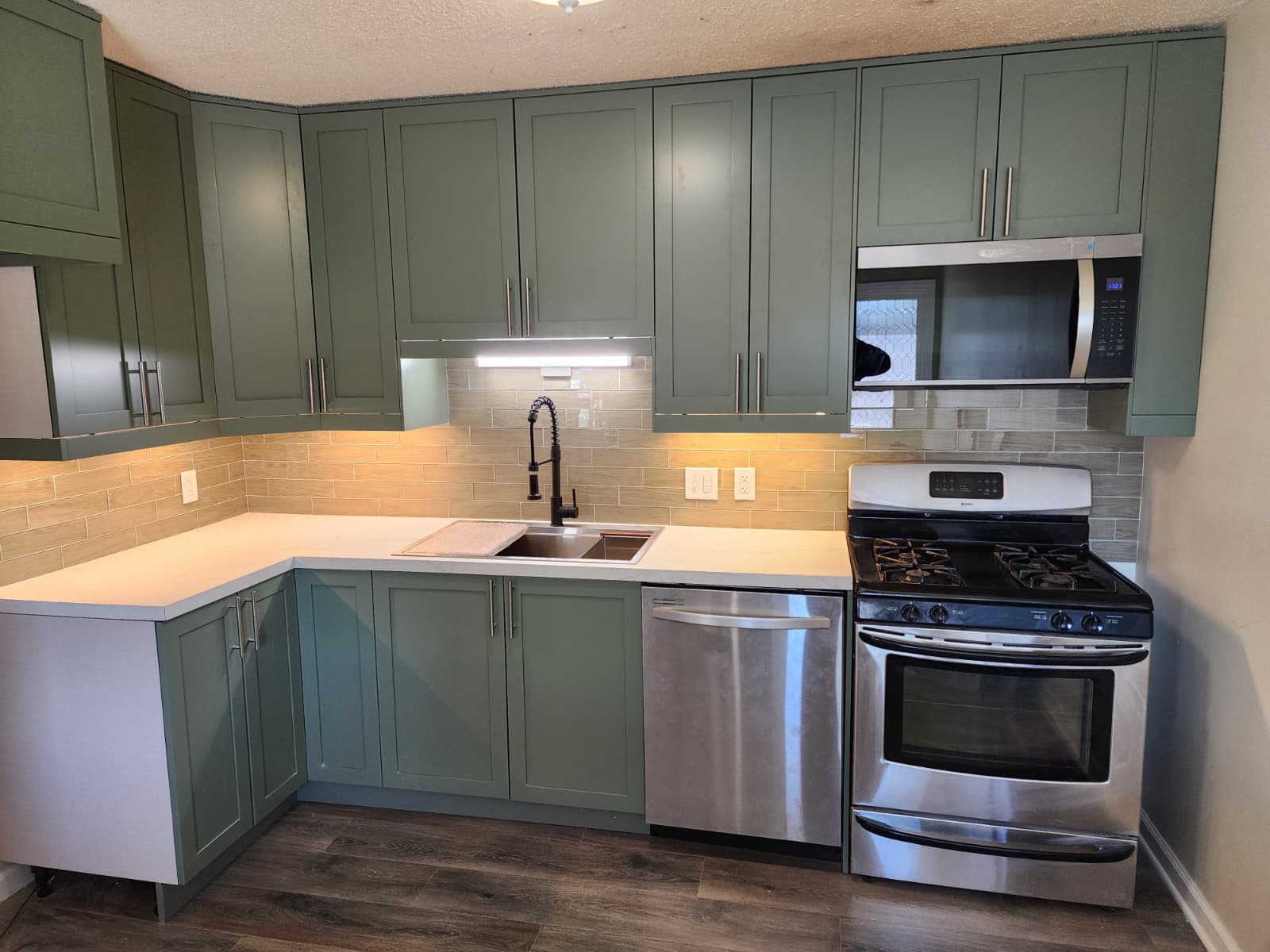 Remodeled kitchen with green shaker cabinets, white quartz counters, tile backsplash, and stainless appliances.